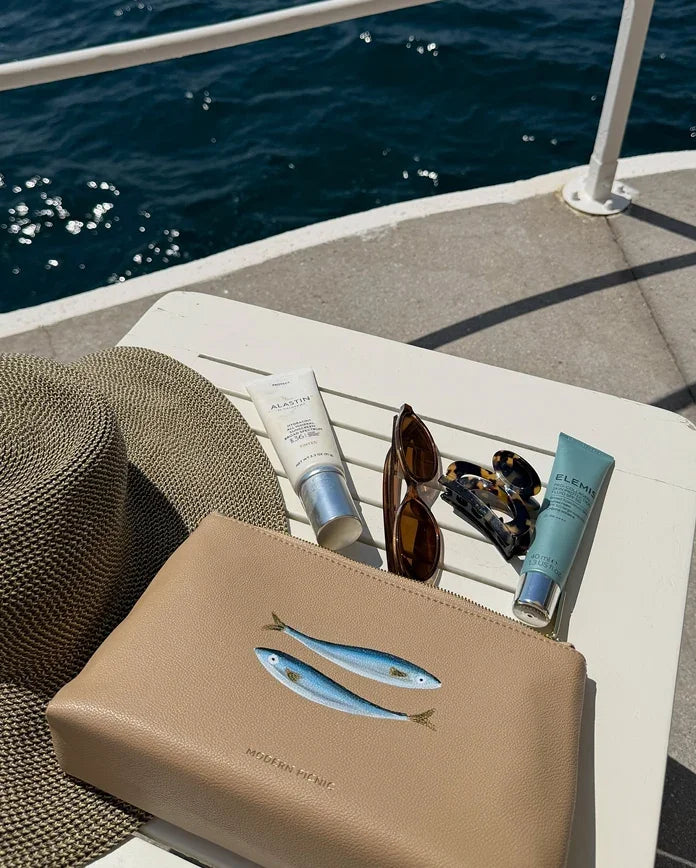 Beige vegan leather clutch with embroidered fish, sunglasses, sunscreen, and hat on boat deck near ocean