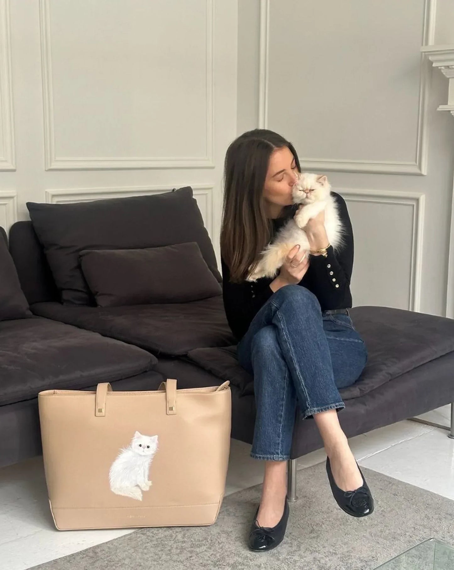 Woman holding fluffy white cat beside beige tote with cat design, styled for everyday chic