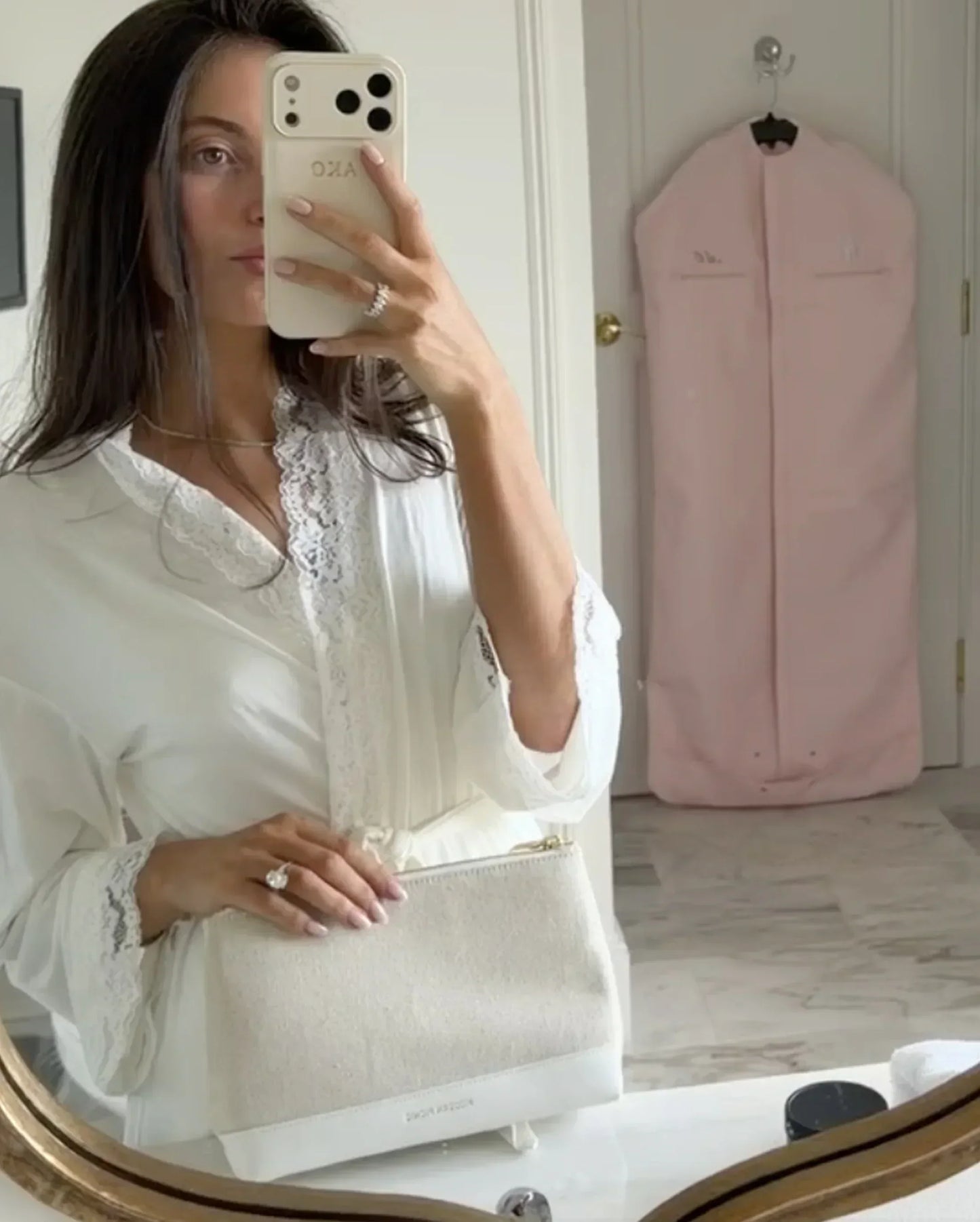 Woman holding chic white vegan leather clutch, taking mirror selfie in lace-trimmed blouse