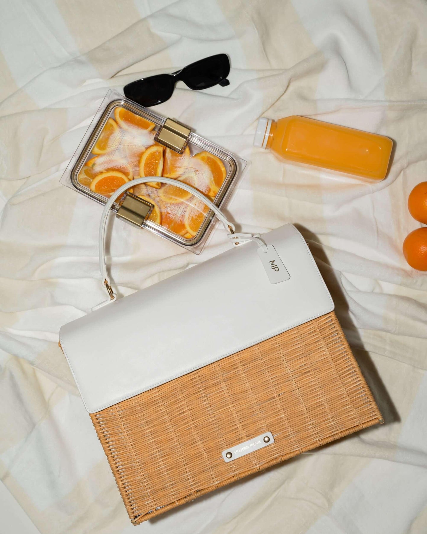 Stylish white and woven picnic tote with orange slices, juice, and sunglasses on a blanket