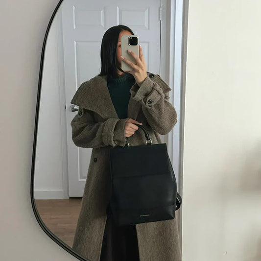 Woman in a brown coat holding a black vegan leather backpack, taking a mirror selfie