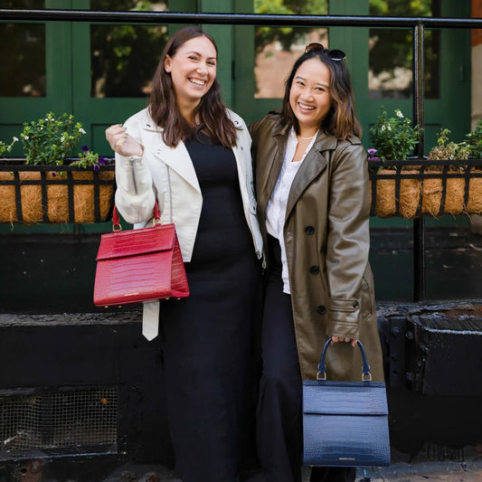 Two women smiling outdoors holding stylish insulated lunch bags, one red and one navy blue.
