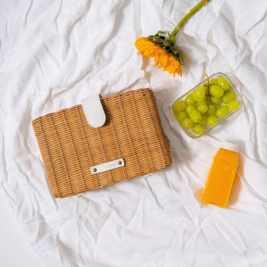 Wicker insulated lunch bag with grapes, orange juice, and a sunflower on a white blanket