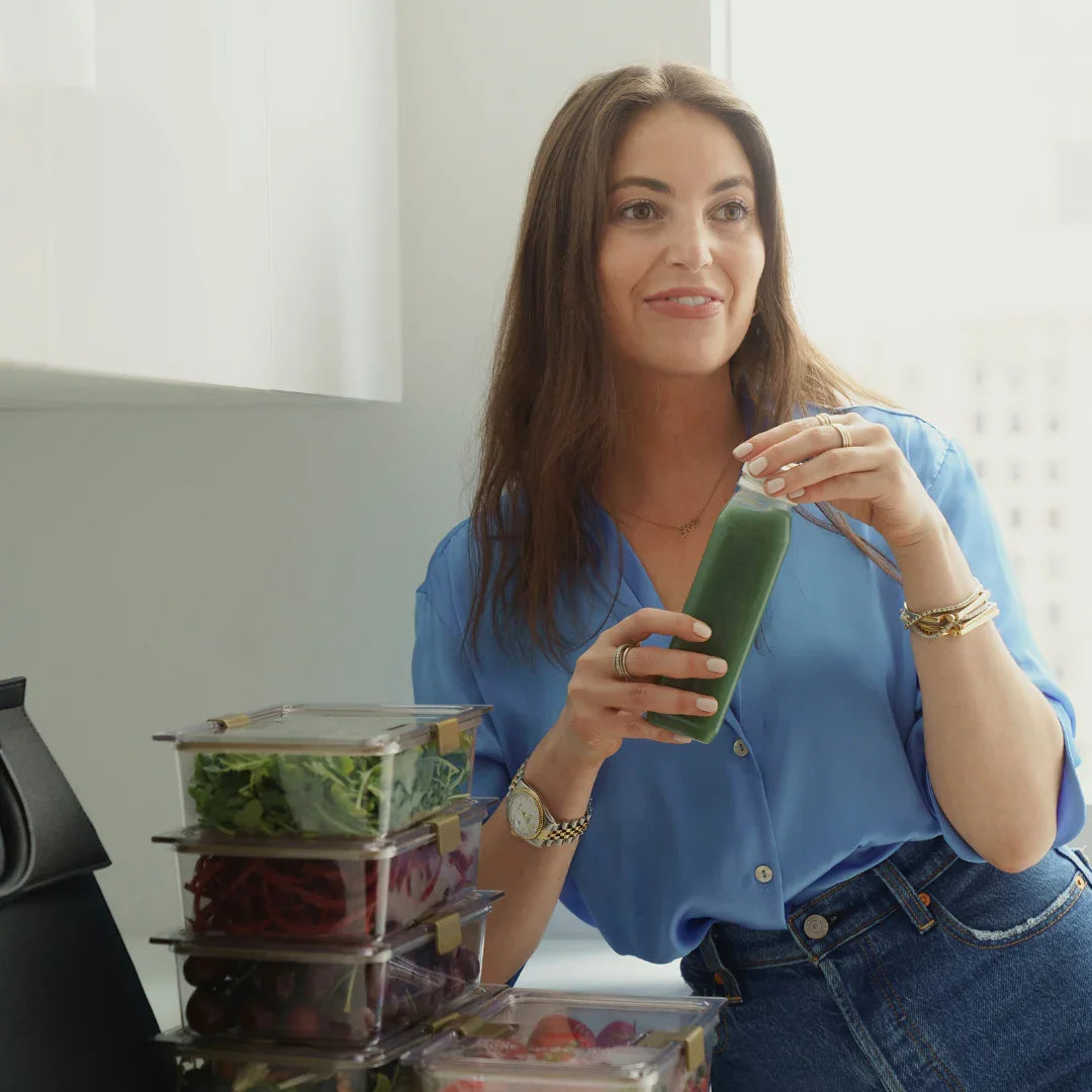 Woman holding green juice with meal prep containers, stylish lunch bag nearby for healthy on-the-go meals