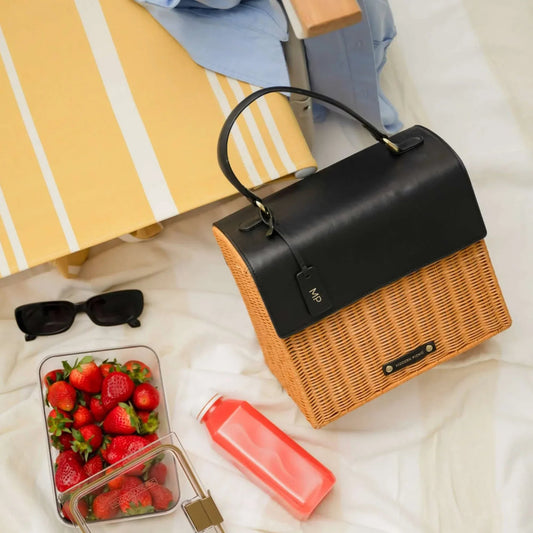 Stylish wicker and black vegan leather lunch tote with strawberries, juice, and sunglasses on picnic blanket