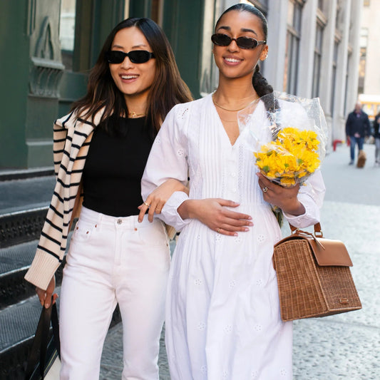 Two women walking city street, one holding yellow flowers and woven tote bag, stylish everyday carry