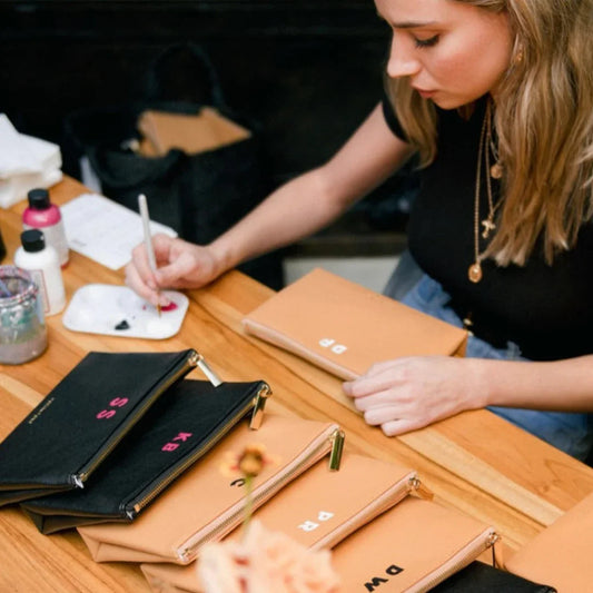 Woman hand-painting initials on tan and black vegan leather pouch bags at a wooden table.