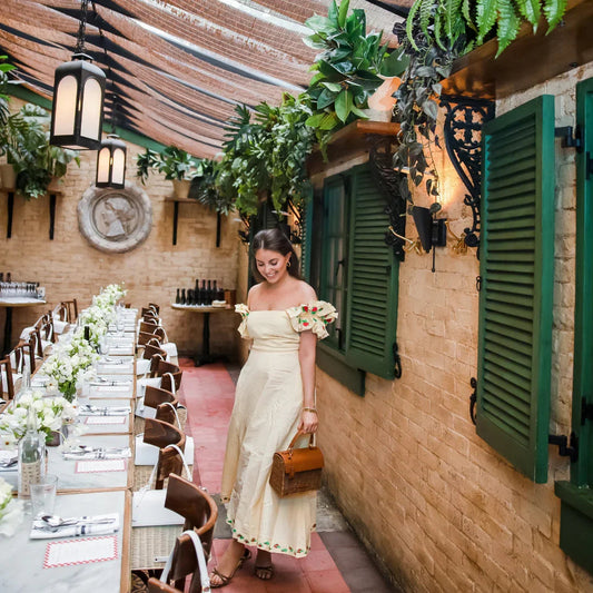 Woman in off-shoulder dress carrying chic woven lunch tote through outdoor dining alley with green shutters