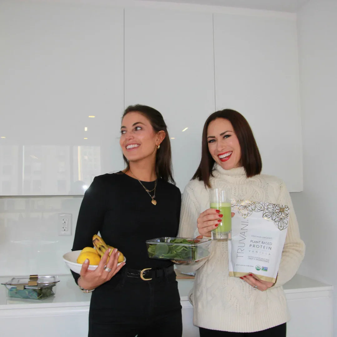 Two women holding fresh bananas, greens, plant-based protein powder, and a green smoothie in a modern kitchen