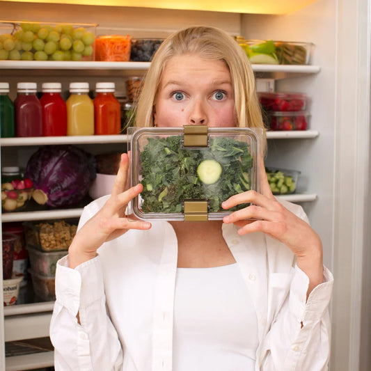 Woman holding clear lunch container with kale salad in front of open fridge, promoting healthy meal prep
