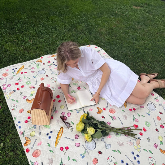 Woman writing on picnic blanket with insulated lunch bag, yellow roses, and sunglasses outdoors