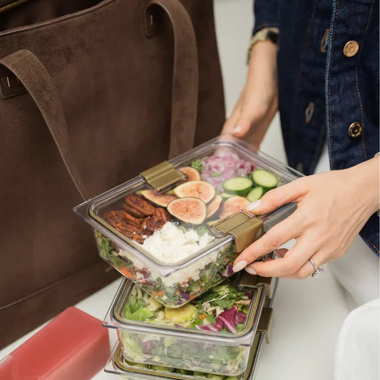 Stacked clear meal prep containers with fresh salad, figs, and nuts, held near a brown tote bag.