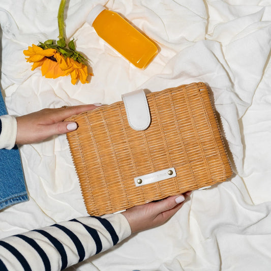 Woven wicker lunch bag with white strap, held on picnic blanket with sunflower and orange juice