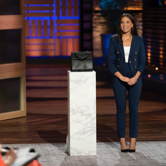 Woman in blue suit presenting a black vegan leather lunch bag on marble pedestal indoors.