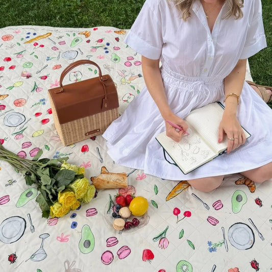 Woman sketching on picnic blanket with insulated lunch bag, fresh fruit, and yellow roses