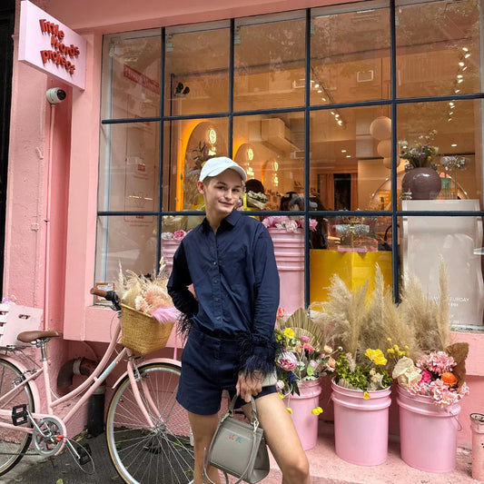 Woman holding vegan leather tote bag in front of pink storefront with flowers and bicycle