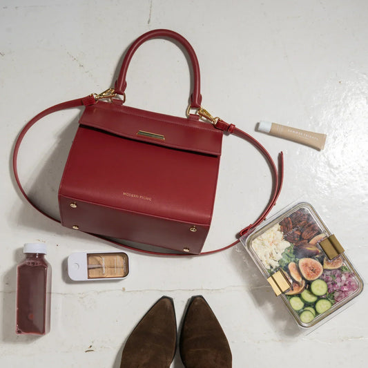 Red insulated lunch bag with handle, salad container, drink, and brown flats on white floor