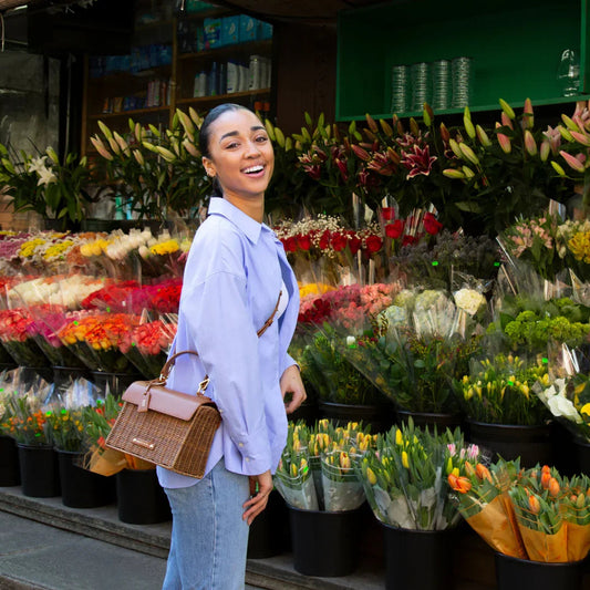 Woman smiling with woven vegan leather tote bag at vibrant flower market, stylish everyday accessory