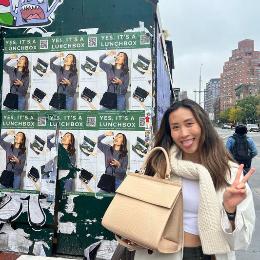 Woman holding beige vegan leather lunch bag, smiling with peace sign near city street ads for insulated lunchbox totes