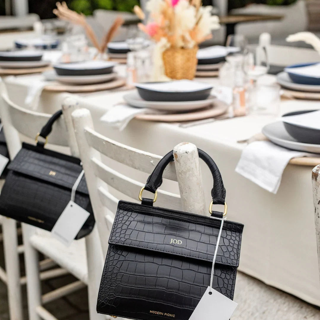 Black croc-texture vegan leather handbag with gold hardware, styled on white chair at outdoor picnic table setting
