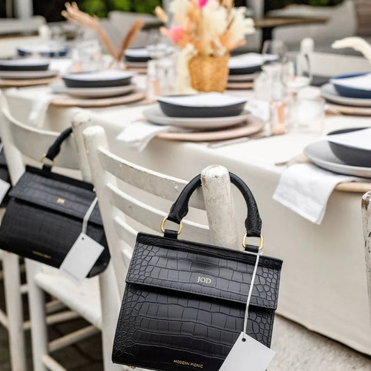Black croc-texture vegan leather handbag with gold hardware, styled on white chair at outdoor picnic table setting