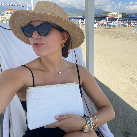 Woman at beach holding white croc-texture clutch, wearing straw hat and sunglasses