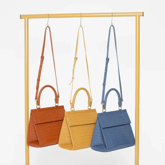 Three croc-texture vegan leather totes in rust, mustard, and blue, hanging on a gold rack for work or travel.
