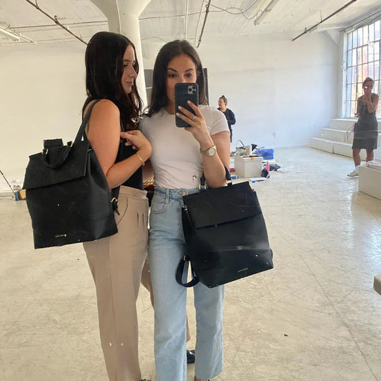 Two women holding black vegan leather backpack totes in a bright studio, perfect for work or travel.