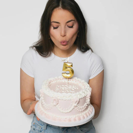 Woman in white shirt holding pink birthday cake with gold number 5 candle and white frosting