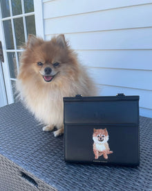 Fluffy tan Pomeranian dog next to black insulated lunch bag with dog illustration, outdoors.