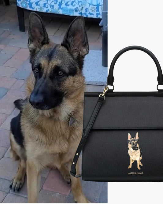 German Shepherd sitting outdoors next to a black vegan leather lunch bag with dog illustration