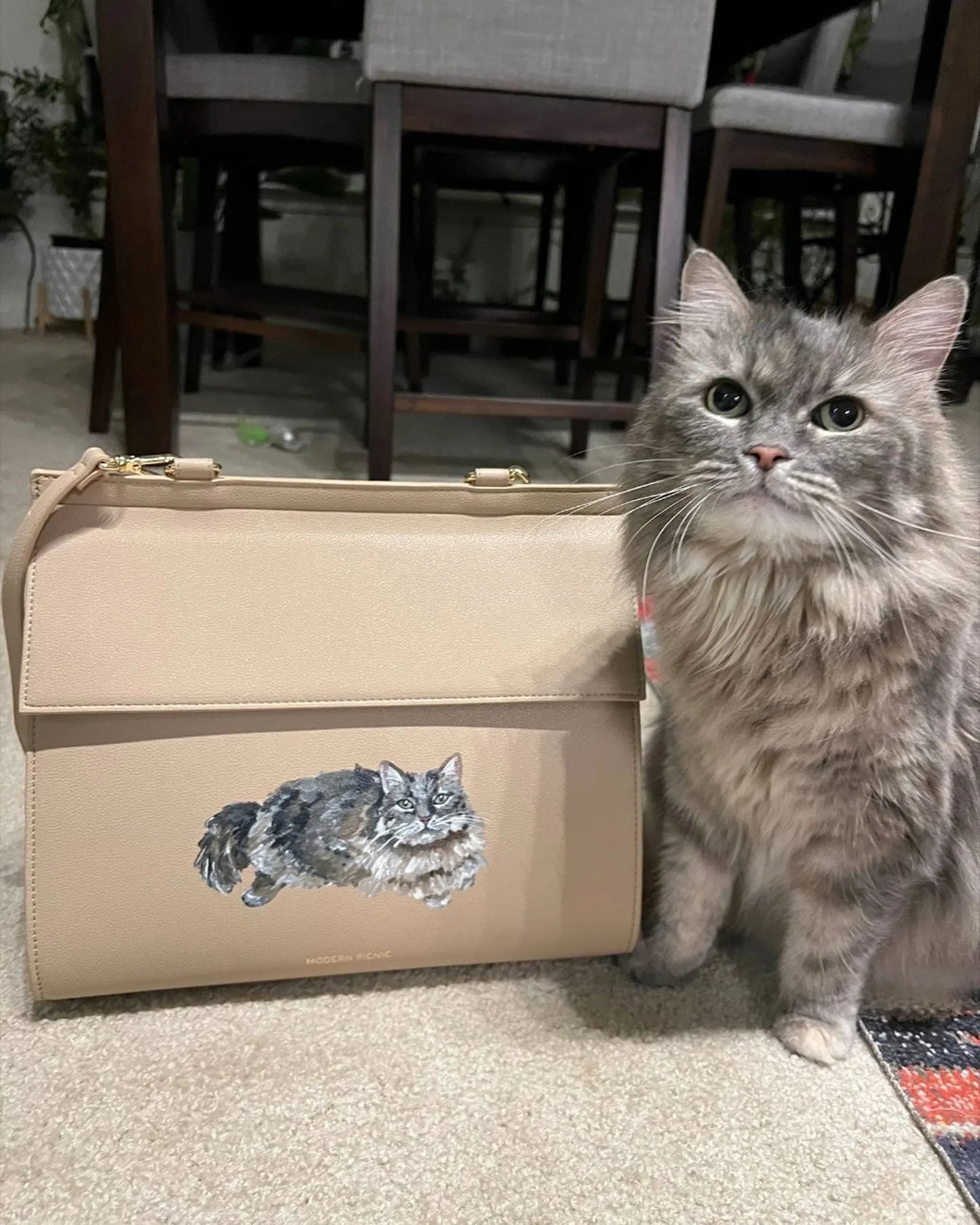Gray fluffy cat next to beige vegan leather lunch bag with cat illustration, indoor setting