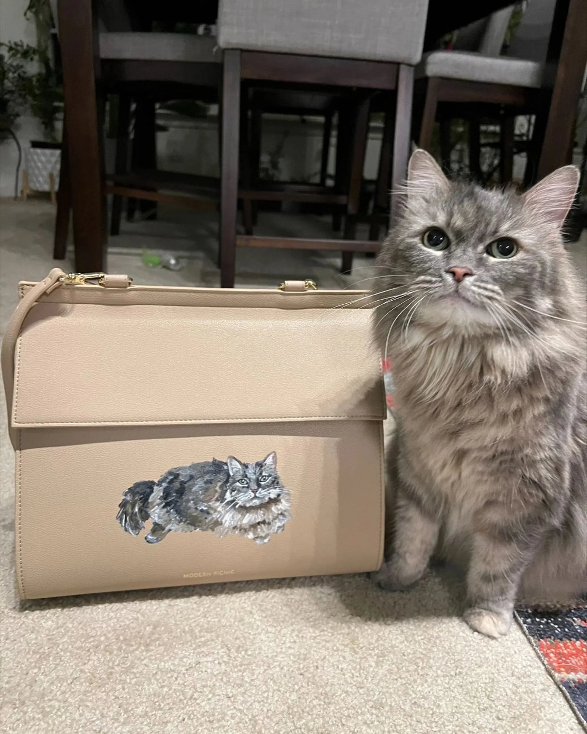 Gray fluffy cat next to beige vegan leather lunch bag with cat illustration, indoor setting