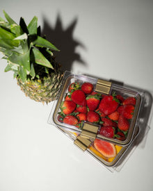 An overhead, angled shot shows two clear, rectangular food containers with gold clips sitting next to a whole fresh pineapple. The top container is filled with strawberries, and the bottom container contains sliced fruit, including papaya.