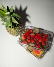 An overhead, angled shot shows two clear, rectangular food containers with gold clips sitting next to a whole fresh pineapple. The top container is filled with strawberries, and the bottom container contains sliced fruit, including papaya.