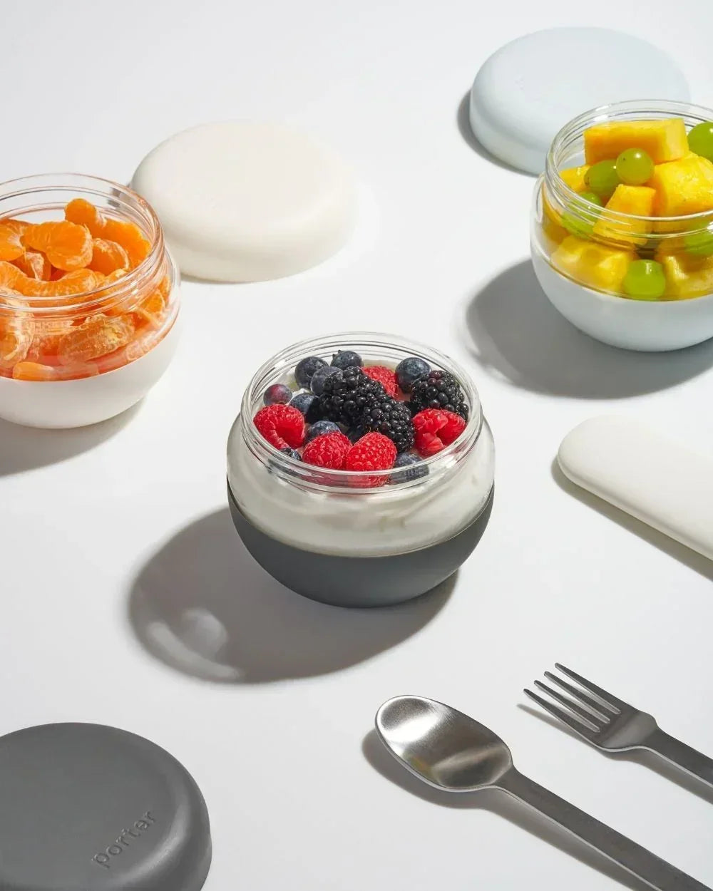 Reusable glass lunch containers with fresh fruit and yogurt on a white table, next to utensils