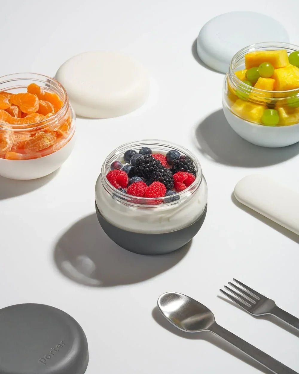 Assorted fruit and yogurt in modern glass lunch containers with cutlery on white background