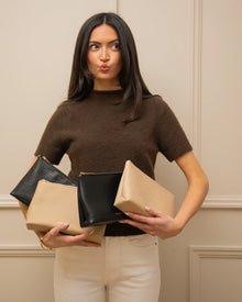Woman holding four vegan leather cosmetic pouches in black and beige, against neutral wall.