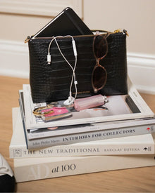 Black vegan leather lunch bag with gold zipper, sunglasses, notebook, and accessories on stacked books