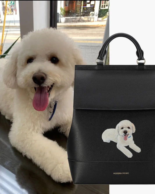 Chic black vegan leather lunch bag with embroidered white dog, next to fluffy dog indoors.