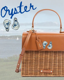 Brown vegan leather picnic bag with oyster design, wicker base, and coastal backdrop
