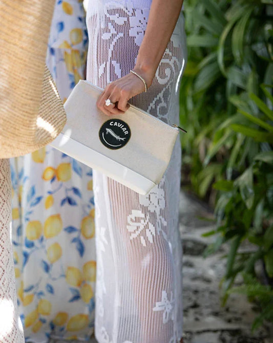 Stylish beige clutch with 'Caviar' logo, held by woman in floral dress, perfect for chic picnics or outings.