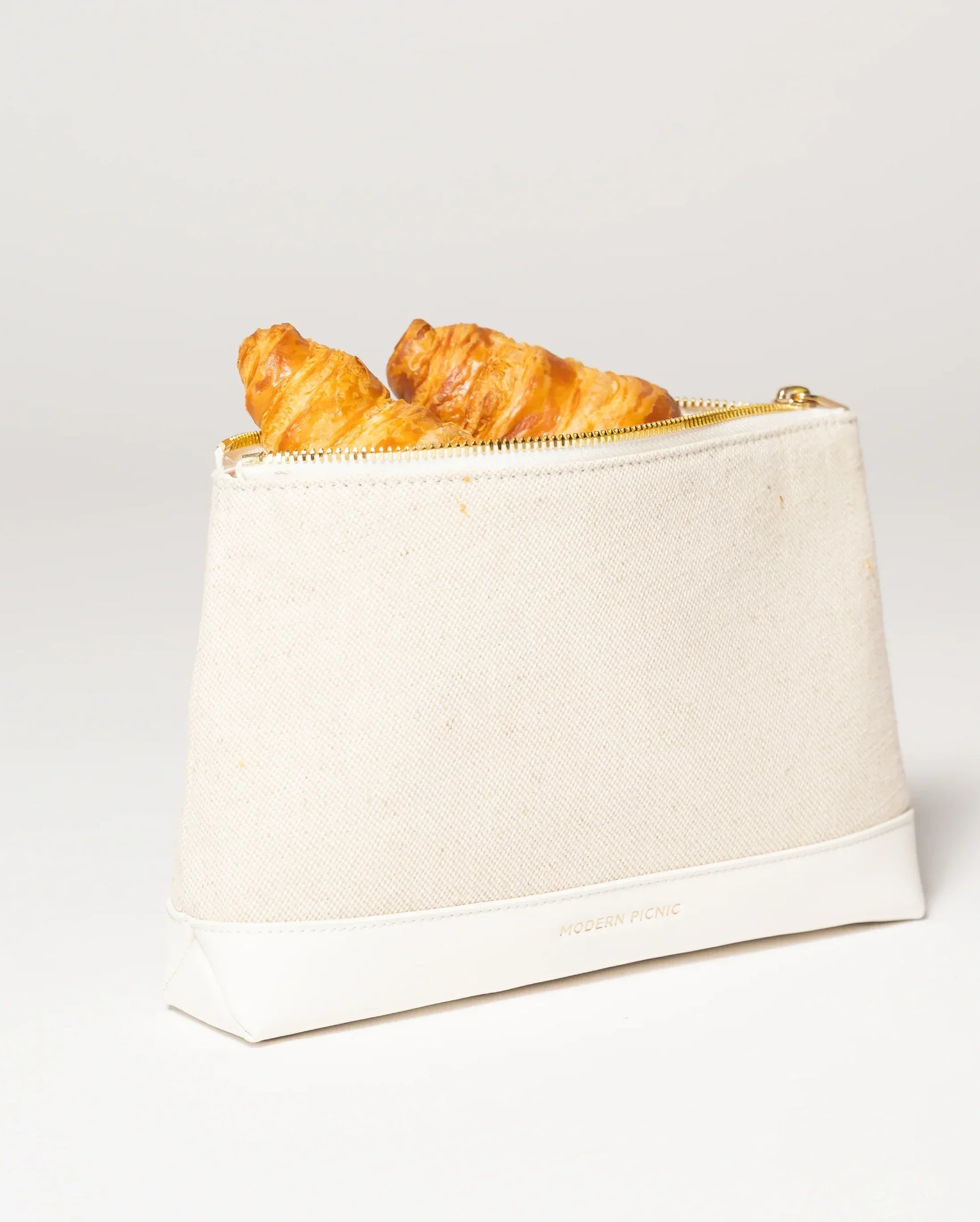 Cream canvas lunch bag with croissants, gold zipper, and vegan leather base for stylish picnics