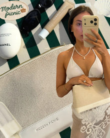 Stylish woman holding a chic beige clutch on a green-and-white striped picnic blanket with beauty essentials.