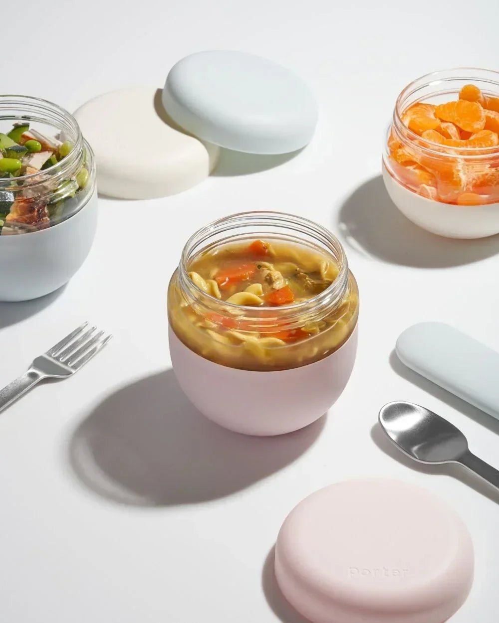 Assorted food in pastel insulated lunch containers with silverware on white background