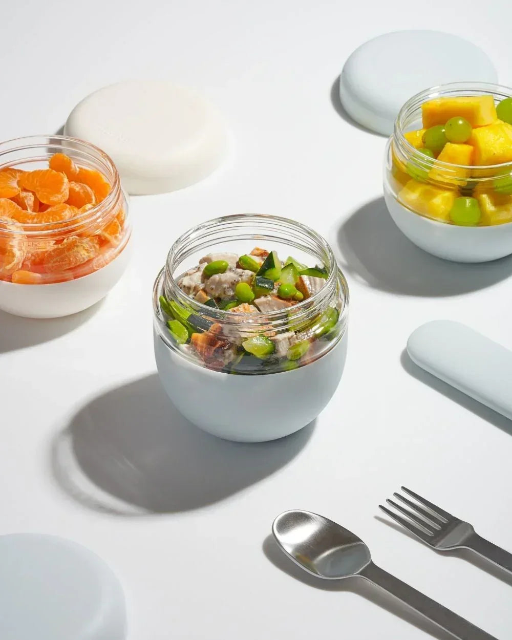 Glass food jars with fruit, salad and stainless steel utensils on a white background