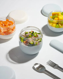 Glass food jars with fruit, salad and stainless steel utensils on a white background