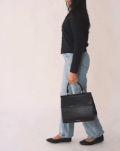 A promotional video still showing a woman holding the black Modern Picnic Large Luncher handbag by its top handle, positioned in the center foreground. She is dressed in a black cardigan, white tank top, and light-wash blue jeans, emphasizing the bag's sleek look as a fashion accessory.