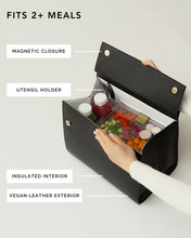 A close-up instructional photo of the open black Modern Picnic Large Luncher handbag. Labels clearly indicate the key features: Vegan Leather Exterior, Insulated Interior, Magnetic Closure, and a slot for a Utensil Holder. The open bag holds a clear food container (salad and vegetables) and two small drink bottles, emphasizing that it FITS 2+ MEALS.