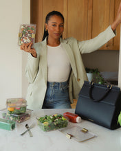 A lifestyle photograph featuring a woman in a light green blazer standing in a bright kitchen. She is surrounded by several clear food containers, including the Modern Picnic Food Container with gold clasps, a juice bottle, and the black Modern Picnic Large Luncher bag, demonstrating the bag's use for chic, organized meal prepping.